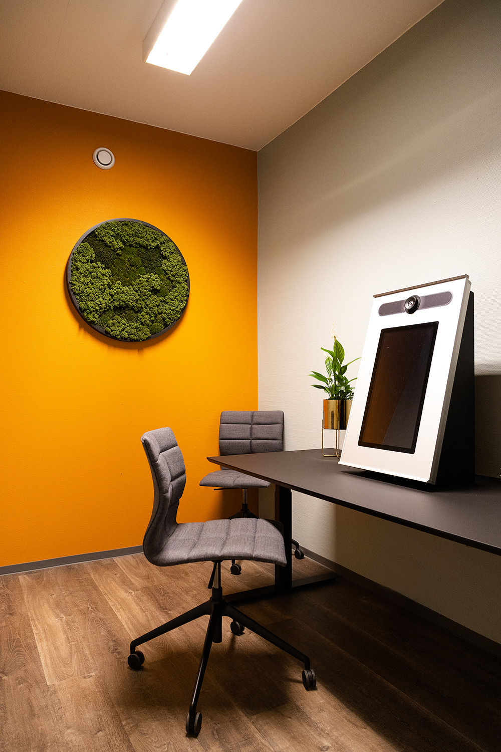Oversiktsbilde av Norges første digitale innbyggertorg som er en rektangulær skjerm med kamera, plassert på høykant. Det sorte bordet står i god kontrast mot den grå veggen, og den oransje veggen i enden av rommet. På den sistnevnte veggen henger en rundt mosekunst. Rommet har to grå stoler og vi ser en plante.