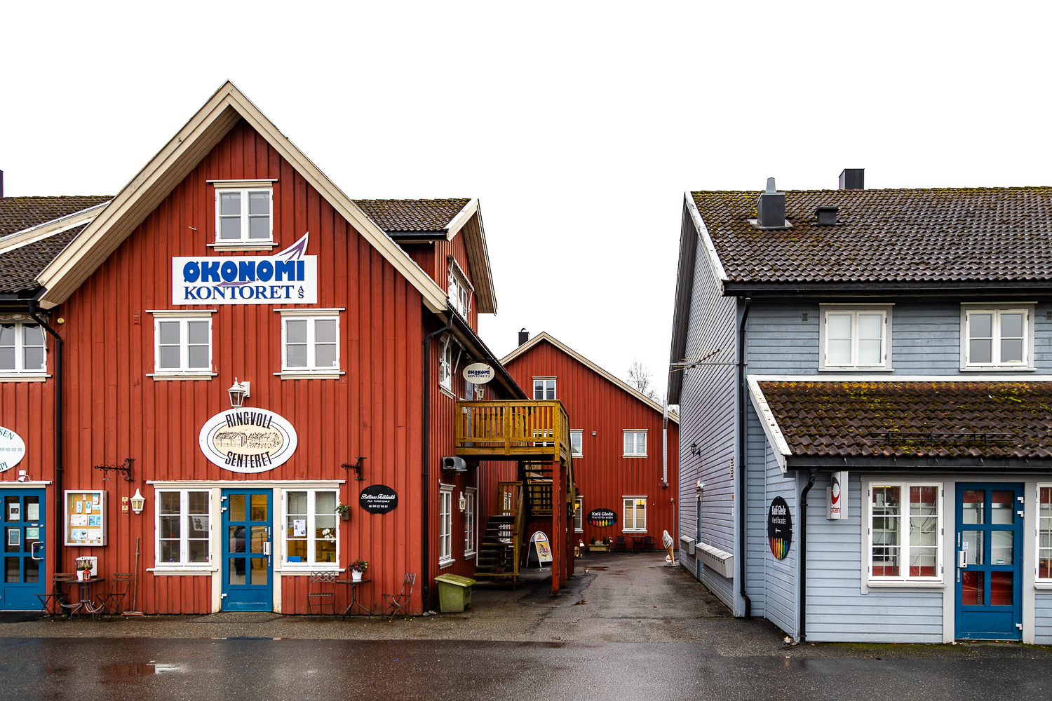 Oversiktsbilde over noe av Ringvoll Senter i Hobøl. Til venstre er et rødt trehus hvor Økonomikontoret AS og Bettinas Fotklinikk holder til, og til høre i det lyseblå trehuset finnes Posten og daglivare.