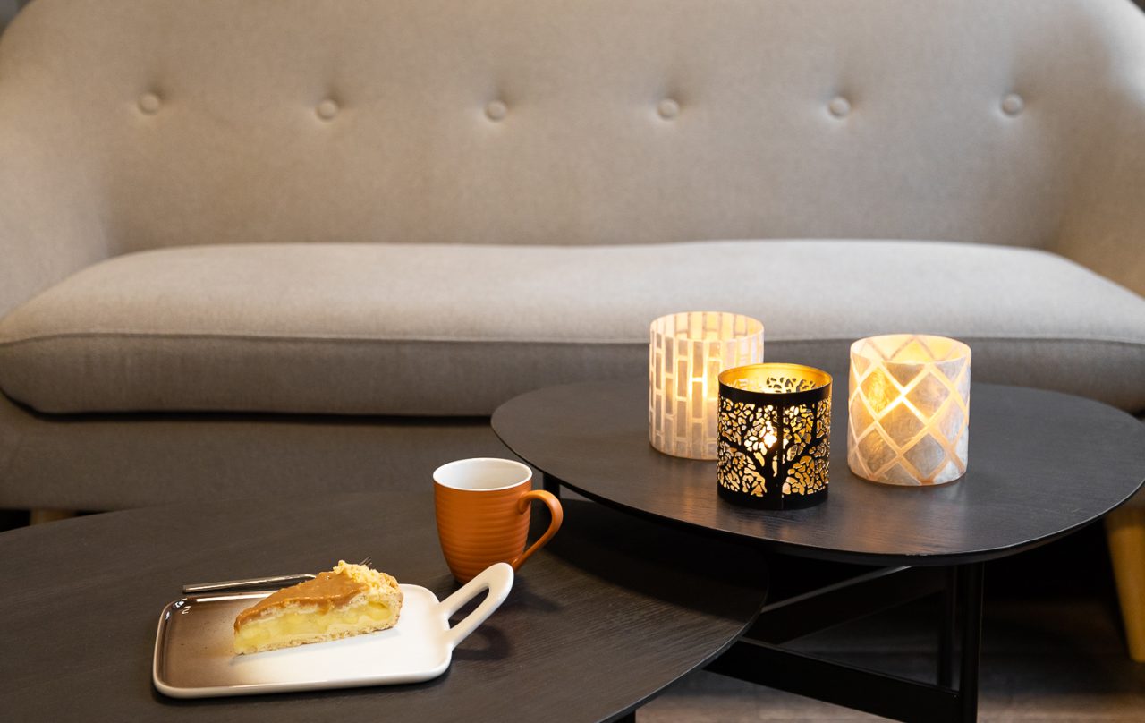 Bilde av et designerfat med kake, en oransje kaffekopp, og tre telysholdere med perlemor og silhutter av trær plassert på små, runde bord. I bakgrunnen skimtes en grå sofa.